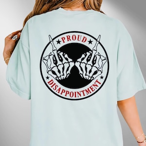May include: Pale blue t-shirt with a black and red graphic on the back. The graphic shows two skeleton hands giving the middle finger, with the words "PROUD" and "DISAPPOINTMENT" around them.