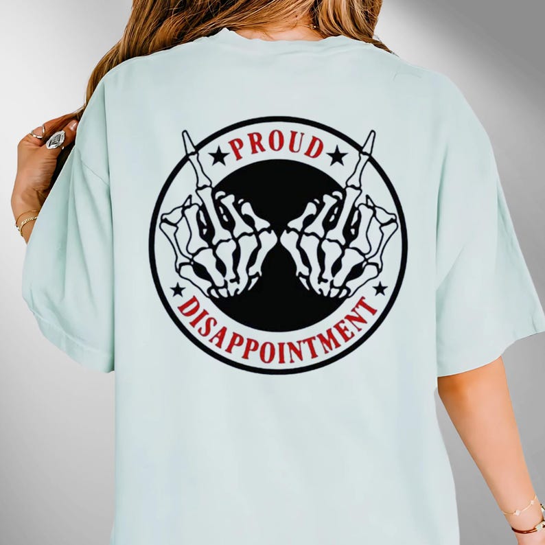 May include: Pale blue t-shirt with a black and red graphic on the back. The graphic shows two skeleton hands giving the middle finger, with the words "PROUD" and "DISAPPOINTMENT" around them.