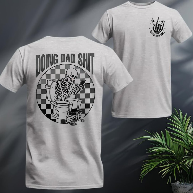May include: Light gray t-shirt with a graphic of a skeleton on a toilet, looking at a phone. The text "DOING DAD SHIT" is above the skeleton. A smaller graphic on the front shows a hand gesture and the same text.