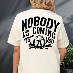 the back of a person wearing a white t-shirt with a black graphic design on it. The design features the text "NOBODY IS COMING TO SAVE YOU" along with a skull and crossbones symbol.