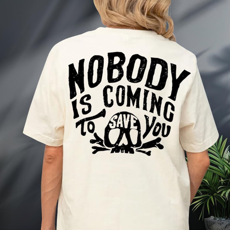 the back of a person wearing a white t-shirt with a black graphic design on it. The design features the text "NOBODY IS COMING TO SAVE YOU" along with a skull and crossbones symbol.