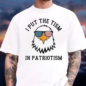 May include: A white short-sleeved t-shirt featuring a graphic of a bald eagle wearing sunglasses with American flag patterns. The text "I PUT THE TISM IN PATRIOTISM" is printed in black capital letters above and below the eagle illustration.