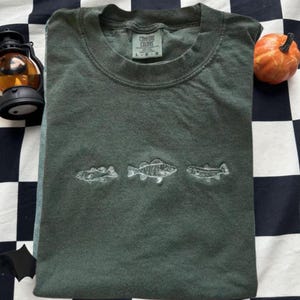 May include: Dark green Comfort Colors t-shirt featuring three embroidered fish designs. The shirt is folded, with a small pumpkin and a lantern in the background. The shirt has a crew neck and short sleeves.