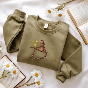 May include: Olive green sweatshirt with a crew neck and long sleeves. The front features an embroidered brown figure with yellow stars. The sweatshirt is displayed on a white surface with daisies and an open book.