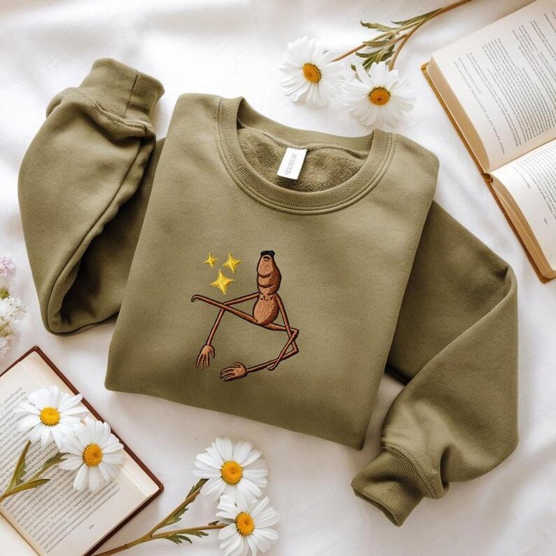 May include: Olive green sweatshirt with a crew neck and long sleeves. The front features an embroidered brown figure with yellow stars. The sweatshirt is displayed on a white surface with daisies and an open book.