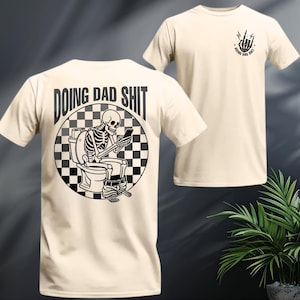 May include: Cream-colored t-shirt featuring a black and white graphic of a skeleton on a toilet, looking at a phone. The text "DOING DAD SHIT" is above the graphic. A smaller graphic with the same text and a hand gesture is on the front.