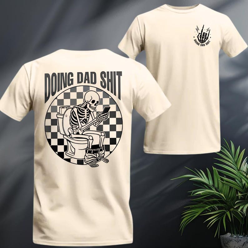 May include: Cream-colored t-shirt featuring a black and white graphic of a skeleton on a toilet, looking at a phone. The text "DOING DAD SHIT" is above the graphic. A smaller graphic with the same text and a hand gesture is on the front.