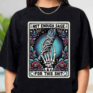 Puede incluir: Camiseta negra de cuello redondo con un dise&ntilde;o gr&aacute;fico. El dise&ntilde;o presenta una mano esquel&eacute;tica sosteniendo un manojo de salvia, rodeada de flores y el texto "NOT ENOUGH SAGE FOR THIS SHIT".
