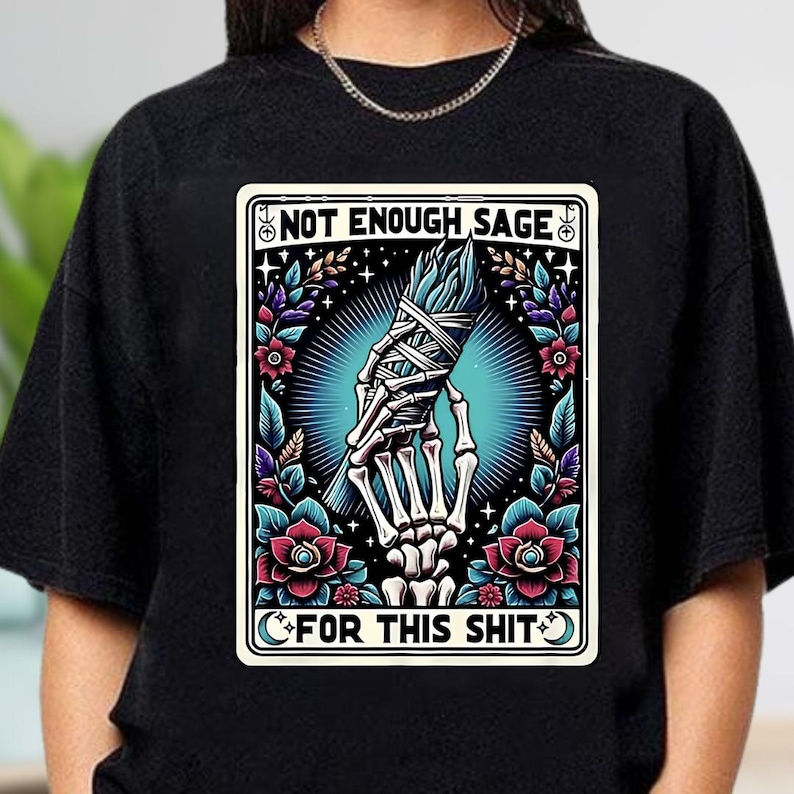 Puede incluir: Camiseta negra de cuello redondo con un dise&ntilde;o gr&aacute;fico. El dise&ntilde;o presenta una mano esquel&eacute;tica sosteniendo un manojo de salvia, rodeada de flores y el texto "NOT ENOUGH SAGE FOR THIS SHIT".