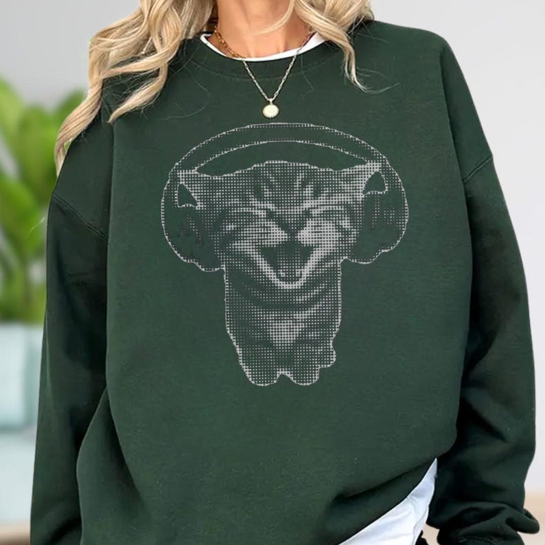 May include: Dark green sweatshirt featuring a pixelated cat wearing headphones. The cat graphic is in shades of gray and white, creating a striking contrast against the solid green background. The sweatshirt has a crew neck and long sleeves, suitable for casual wear.