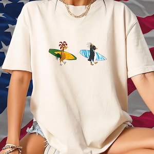 May include: Cream-colored t-shirt featuring cartoon illustrations of a rooster and a penguin, each holding a surfboard. The rooster has a red comb and the penguin has a black head. The surfboards are green and blue.