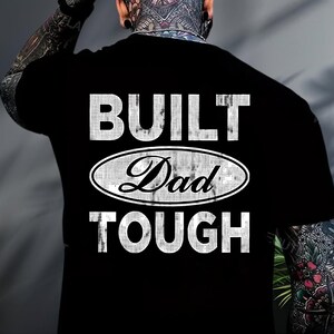 May include: Black t-shirt with the words "BUILT Dad TOUGH" in white, distressed lettering. The word "Dad" is inside an oval design. The shirt is viewed from the back.