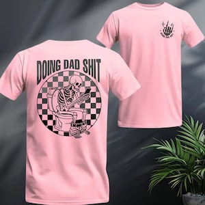 May include: Pink t-shirt featuring a black and white graphic of a skeleton on a toilet, looking at a phone. The text "DOING DAD SHIT" is above the graphic. A smaller graphic with the text "DOING DAD SHIT" and a hand gesture is on the front.