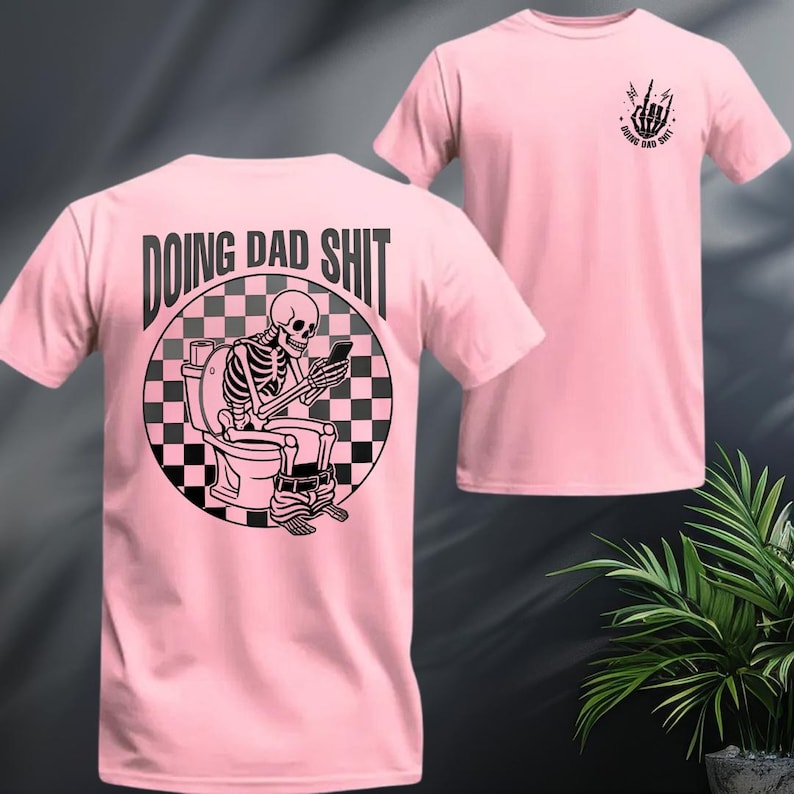 May include: Pink t-shirt featuring a black and white graphic of a skeleton on a toilet, looking at a phone. The text "DOING DAD SHIT" is above the graphic. A smaller graphic with the text "DOING DAD SHIT" and a hand gesture is on the front.