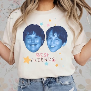 May include: Cream-colored t-shirt featuring two blue-toned faces and the text "BEST FRIENDS" in a playful font. The design includes colorful stars. The shirt has a classic crew neck and short sleeves.