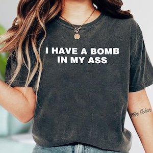 May include: Dark gray t-shirt with the white text "I HAVE A BOMB IN MY ASS". The shirt has a crew neck and short sleeves. A gold necklace is visible at the neckline.