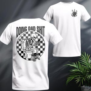 May include: White t-shirt with a black and white graphic of a skeleton on a toilet, looking at a phone. The text "DOING DAD SHIT" is above the graphic. A smaller graphic of a hand gesture and text "DOING DAD SHIT" is on the front.