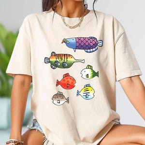 May include: Beige t-shirt featuring a colorful fish graphic. The design includes various cartoon fish in shades of blue, purple, green, red, and yellow. The shirt is a casual, everyday wear item.