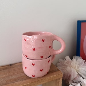 May include: Two stacked handmade ceramic mugs with a light pink glaze and a pattern of small, hand-painted red hearts. The mugs feature an irregular, organic shape with a thick, rounded handle on the top mug and a smooth, glossy finish.
