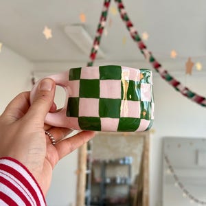 Pode incluir: Uma caneca de cerâmica com acabamento brilhante apresentando um padrão de xadrez pintado à mão em tons alternados de verde floresta e rosa claro. A caneca tem uma forma arredondada e irregular com uma alça curva na lateral.