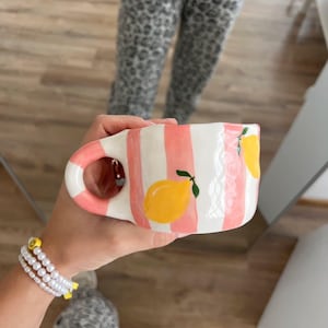 Könnte beinhalten: Eine Keramiktasse mit rosa und weißen Streifen und gelben Zitronenillustrationen. Die Tasse hat einen rosa Griff. Ein Perlenarmband mit einem gelben Smiley-Anhänger ist ebenfalls sichtbar. Die Tasse wird von einer Person gehalten.