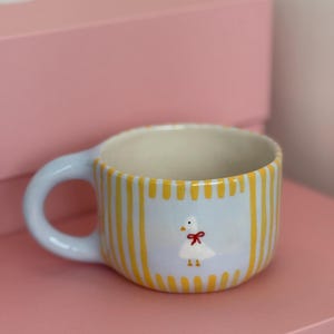 Puede incluir: Una taza de cerámica con asa e interior azul claro. El exterior presenta rayas verticales amarillas y un panel azul claro con un ganso de dibujos animados con un lazo rojo. La taza está sobre una superficie rosa.