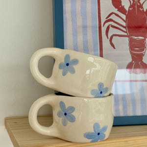 Puede incluir: Dos tazas de cerámica blanquecina apiladas sobre una estantería de madera. Cada taza presenta un asa grande y curvada y está decorada con diseños florales azules. El fondo incluye una impresión enmarcada con una ilustración de langosta.