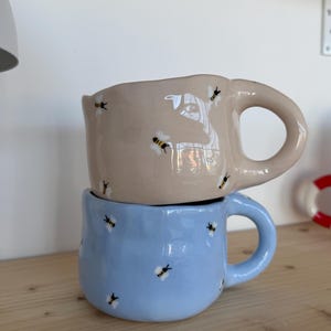 Puede incluir: Dos tazas de cerámica apiladas en una estantería de madera. La taza superior es beige, la inferior azul claro. Ambas tazas tienen un asa redondeada y están decoradas con pequeñas ilustraciones de abejas. Parte de una colección de utensilios de cocina.