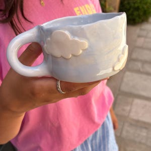 Puede incluir: Una taza de cerámica azul claro con acabado brillante y borde irregular y ondulado. La taza presenta apliques de nubes blancas en relieve tridimensional en la superficie exterior y un asa curva en el lado izquierdo para sujetar el recipiente.