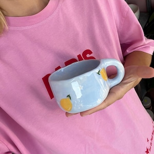 Könnte beinhalten: Eine hellblaue Keramiktasse mit weißem Überzug und einem gelben Zitronenmotiv. Die Tasse hat eine abgerundete Form und einen Henkel.