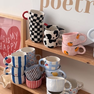 May include: A collection of ceramic mugs with various hand-painted patterns including black and white checkerboard, cow print, pink with orange fruit, blue stripes with red hearts, blue and white gingham with cherries, and red or brown vertical stripes.