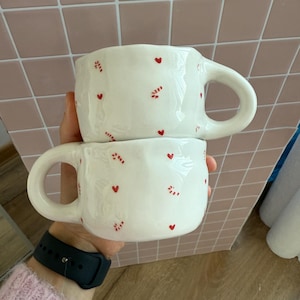 Può includere: Due tazze in ceramica bianca con una texture ondulata e grandi manici. Ogni tazza è decorata con piccoli cuori rossi e gruppi di punti rossi. Le tazze sono impilate l'una sull'altra.