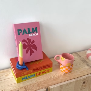May include: A stack of colorful books with the text "PALM BEACH", "CAPRI DOLCE VITA", and "MIAMI BEACH". A pink mug with a smiley face design sits next to the books. A blue candle holder and a two-tone candle are also present.