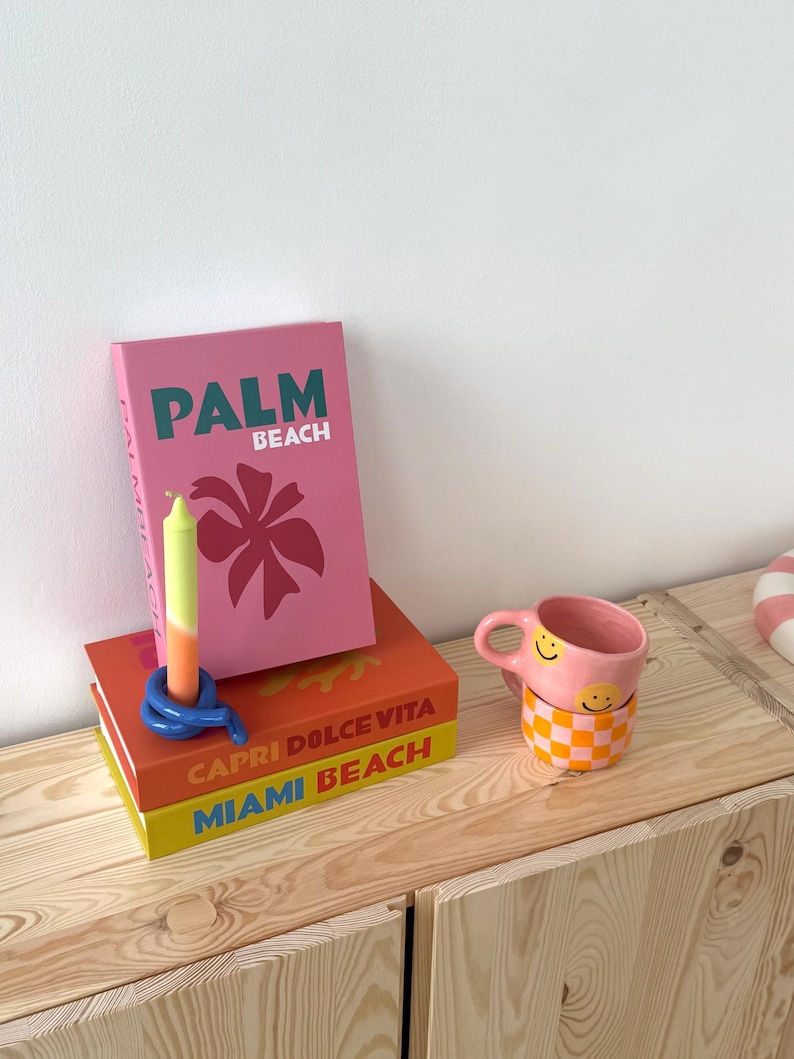 May include: A stack of colorful books with the text "PALM BEACH", "CAPRI DOLCE VITA", and "MIAMI BEACH". A pink mug with a smiley face design sits next to the books. A blue candle holder and a two-tone candle are also present.