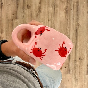 May include: A light pink ceramic mug featuring a repeating pattern of small, bright red crabs with black eyes and white polka dots scattered across the surface. The mug has a rounded, integrated handle and a smooth, glossy finish throughout.
