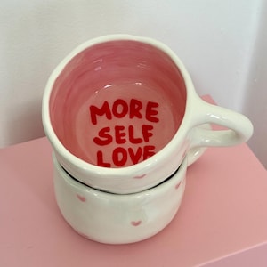 Peut inclure: Mug en c&eacute;ramique blanche avec int&eacute;rieur rose et l'inscription "MORE SELF LOVE" en rouge. La tasse a une anse et est empil&eacute;e sur un bol assorti d&eacute;cor&eacute; de petits c&oelig;urs roses. L'ensemble est pos&eacute; sur une surface rose.