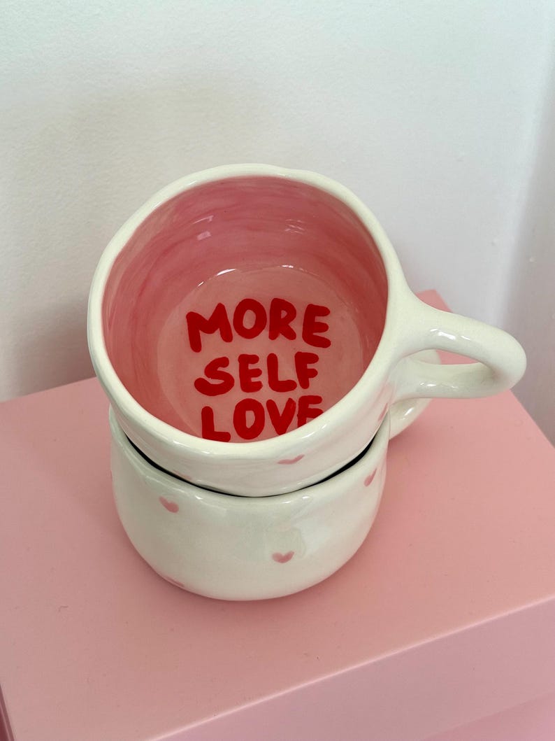 Peut inclure: Mug en c&eacute;ramique blanche avec int&eacute;rieur rose et l'inscription "MORE SELF LOVE" en rouge. La tasse a une anse et est empil&eacute;e sur un bol assorti d&eacute;cor&eacute; de petits c&oelig;urs roses. L'ensemble est pos&eacute; sur une surface rose.