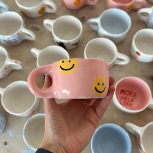 May include: A collection of handmade ceramic mugs in various colors and designs. A pink mug with two yellow smiley faces is held in the foreground. Other mugs include blue, white, and pink with patterns and the text "MORE SELF LOVE".