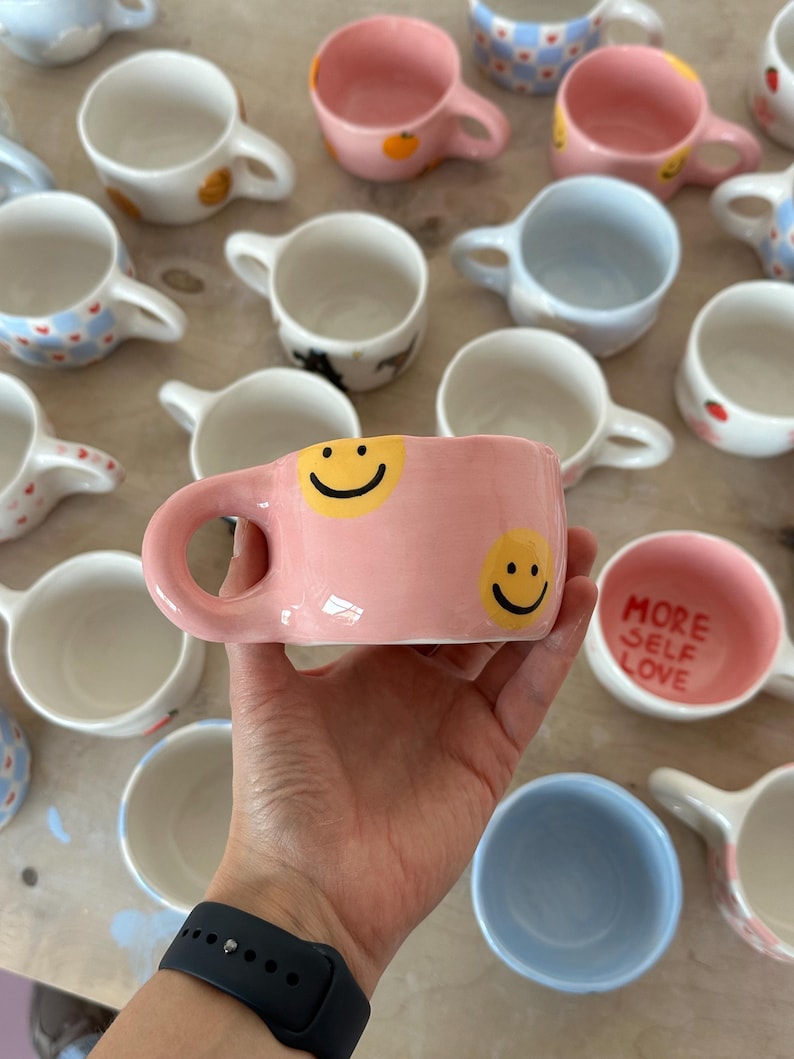 May include: A collection of handmade ceramic mugs in various colors and designs. A pink mug with two yellow smiley faces is held in the foreground. Other mugs include blue, white, and pink with patterns and the text "MORE SELF LOVE".