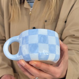 May include: A ceramic mug with a light blue and white chequered pattern. The mug has a rounded handle and a square shape. The mug is held in a person's hands.
