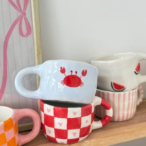 Può includere: Una collezione di tazze in ceramica con design unici. Una tazza presenta un granchio rosso su sfondo azzurro. Un'altra ha un motivo a scacchi rosso e bianco con cuori rosa. Altre tazze hanno disegni di anguria e a righe.
