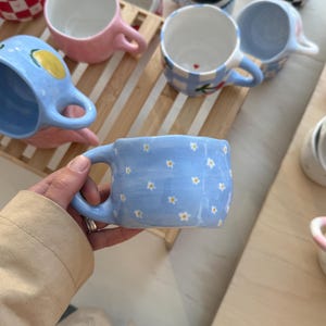 May include: A light blue ceramic mug decorated with small white daisy flowers. The mug has a curved handle. Other mugs with different patterns and colours are visible in the background.