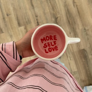 Peut inclure: Une tasse en c&eacute;ramique blanche avec un int&eacute;rieur rose et l'inscription "MORE SELF LOVE" en rouge. La tasse a une anse blanche. La personne tient la tasse et porte un haut de pyjama rose &agrave; rayures.