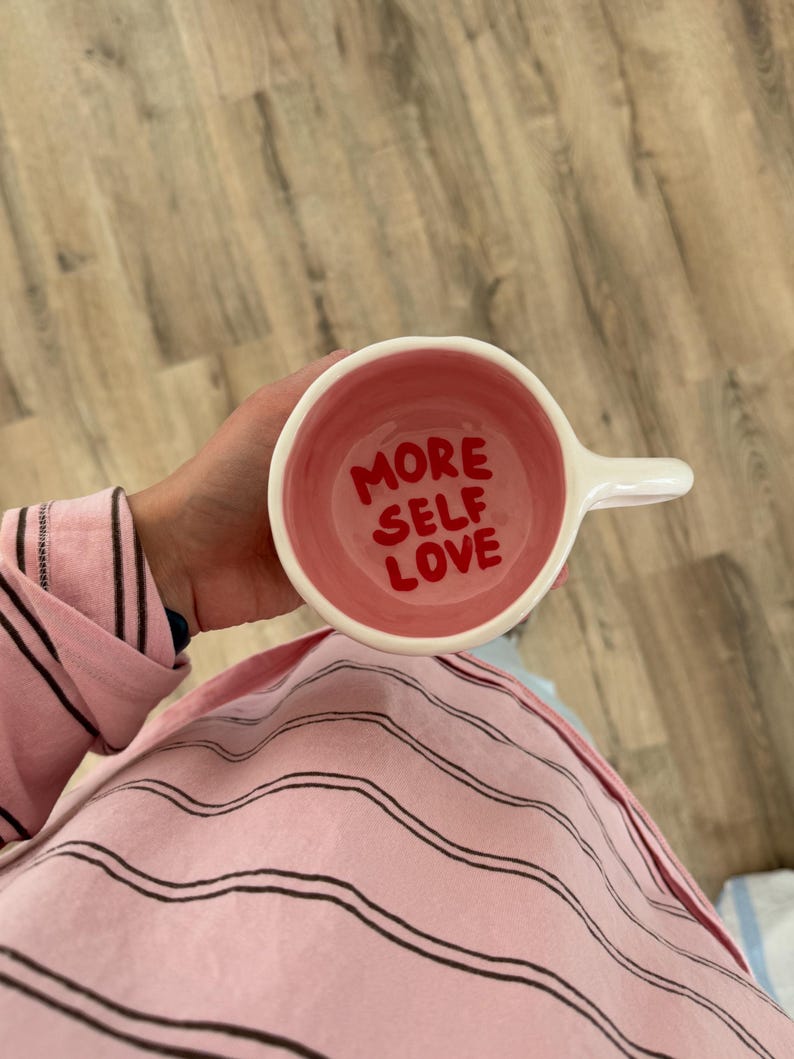 Peut inclure: Une tasse en c&eacute;ramique blanche avec un int&eacute;rieur rose et l'inscription "MORE SELF LOVE" en rouge. La tasse a une anse blanche. La personne tient la tasse et porte un haut de pyjama rose &agrave; rayures.