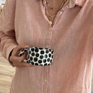 Puede incluir: Una taza de cerámica blanca con un patrón de lunares negros se sostiene frente a una camisa rosa abotonada. La taza tiene un asa curva y está llena de un líquido desconocido. La camisa es de color rojo claro.