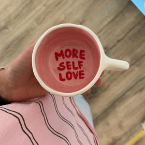May include: White ceramic mug with a pink interior. The words "MORE SELF LOVE" are written in red inside the mug. The mug has a handle and is held by a person.