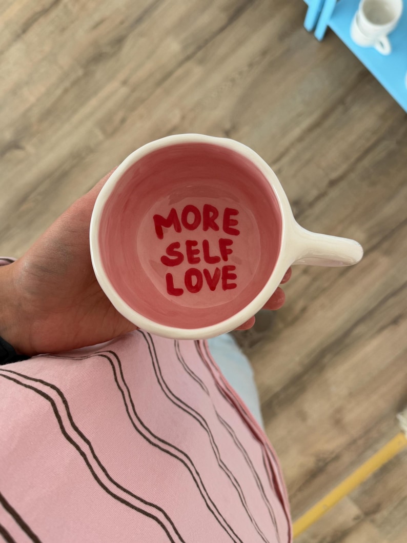 Peut inclure: Tasse en c&eacute;ramique blanche avec un int&eacute;rieur rose. Les mots "MORE SELF LOVE" sont &eacute;crits en rouge &agrave; l'int&eacute;rieur de la tasse. La tasse a une anse et est tenue par une personne.
