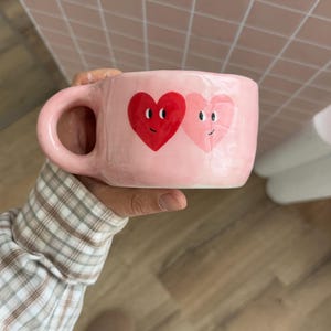 Pode incluir: Uma caneca de cerâmica rosa com uma alça grande, com dois corações de desenho animado com rostos sorridentes. Um coração é vermelho, o outro rosa. A caneca é segurada por uma pessoa.