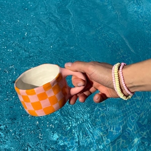 Puede incluir: Una taza de cerámica a cuadros rosa y naranja con asa. La taza se sostiene sobre una piscina azul.