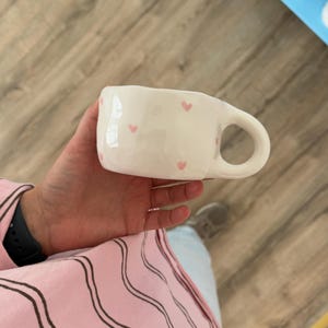 Peut inclure: Une tasse en c&eacute;ramique blanche avec une anse arrondie et des motifs en forme de c&oelig;ur roses. La tasse est tenue dans la main d'une personne. L'arri&egrave;re-plan comprend un sol en bois et un aper&ccedil;u d'une &eacute;tag&egrave;re bleue.